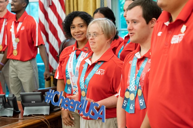 Special olympics team wearing red shirts with a woman with a medal around her neck. In her hand is a sign that reads "#cheer4USA"