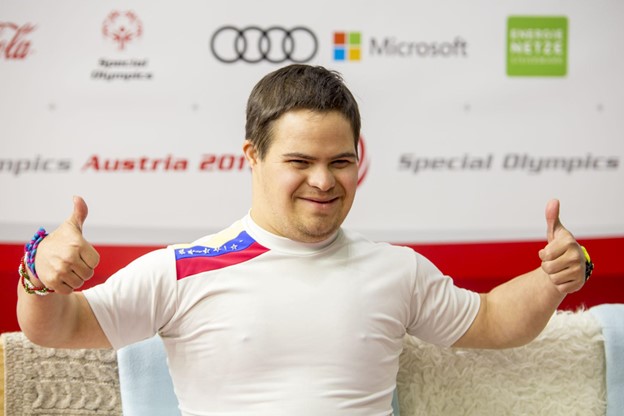 Man with disability in white shirt at special olympics smiling with two thumbs up.