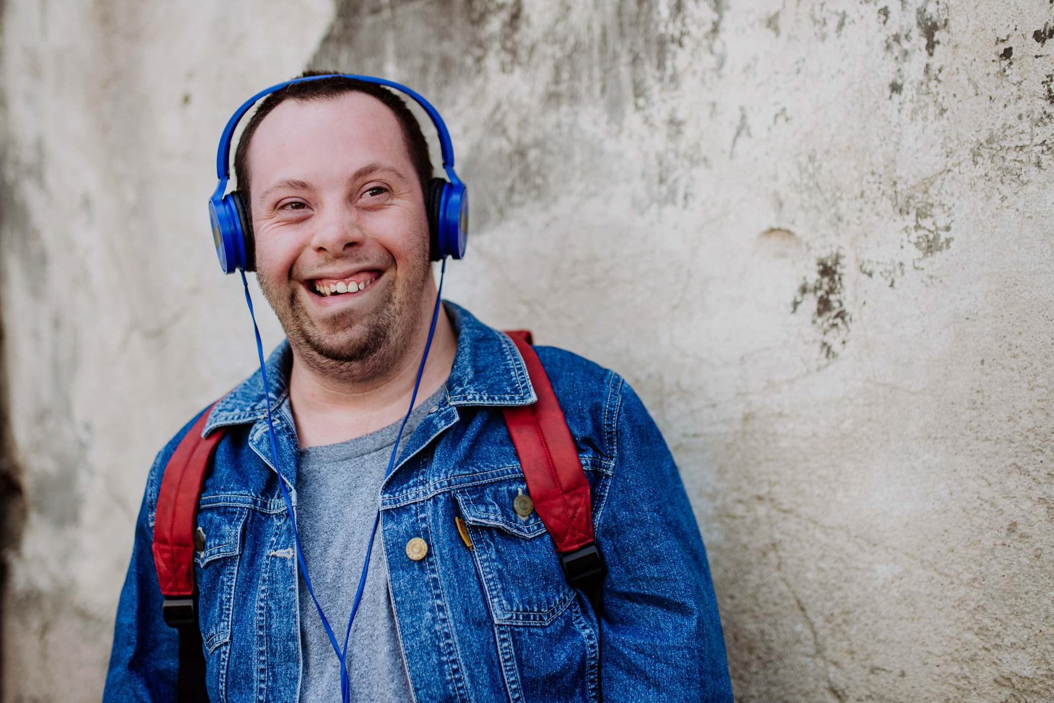 Close-up portrait of happy young man with Down sydrome listening to music when walking in street. Man with a disability wearing headphones and jeans jacket