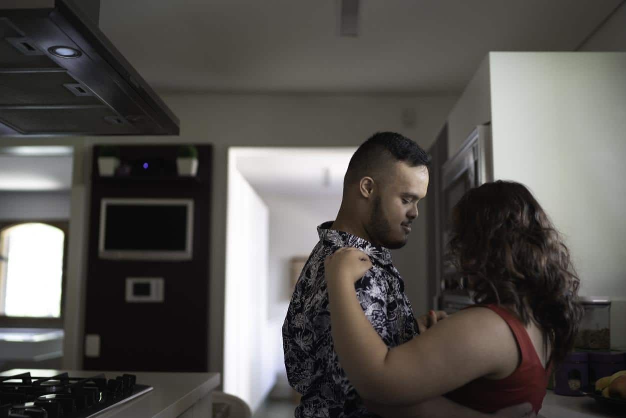 Special needs couple dancing in the kitchen Hands holding open a Book with the 3 icons on a light background, An ear, a person in a wheelchair and eye. On the otherside of the book, on a dark back ground is the test ADA American Disabilities Act.