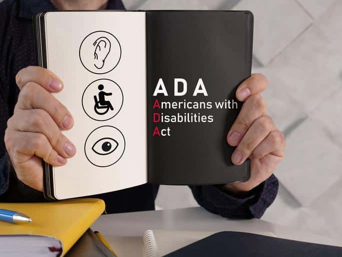 Hands holding open a Book with the 3 icons on a light background, An ear, a person in a wheelchair and eye. On the otherside of the book, on a dark back ground is the test ADA American Disabilities Act.