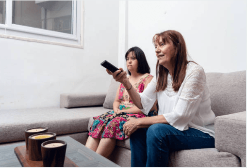 Caregiver and young woman with down syndrome sitting on the couch. Caregiver has remote in her hand