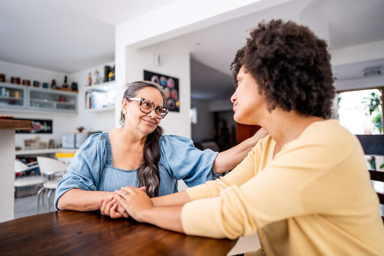 Senior mother consoling her daughter at home Older woman holding the hand of another woman sitting at a table