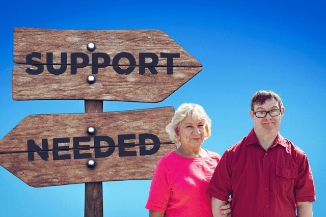 Adult Mom standing with Adult son with a disability in front of a sign that says Support Needed