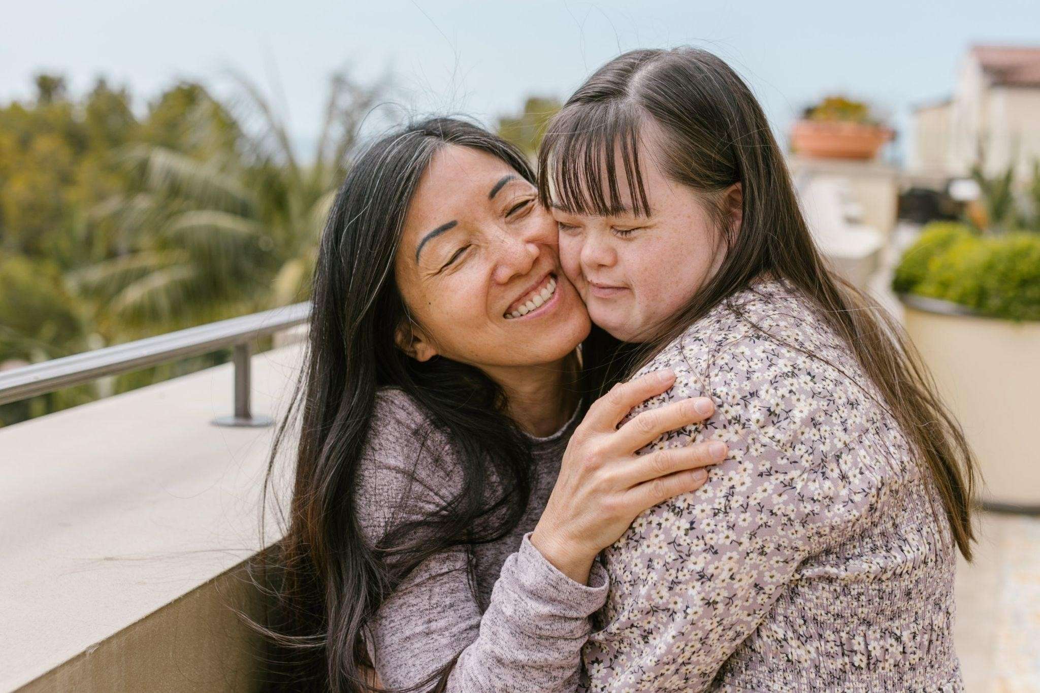 A middle-aged woman smiles and embraces a young woman with down syndrome.