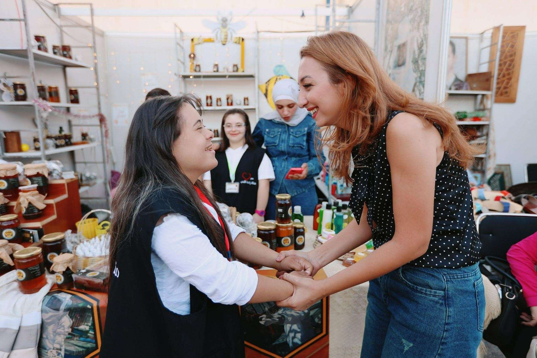 Two women warmly hold hands while in a small shop. Two other women stand in the background.