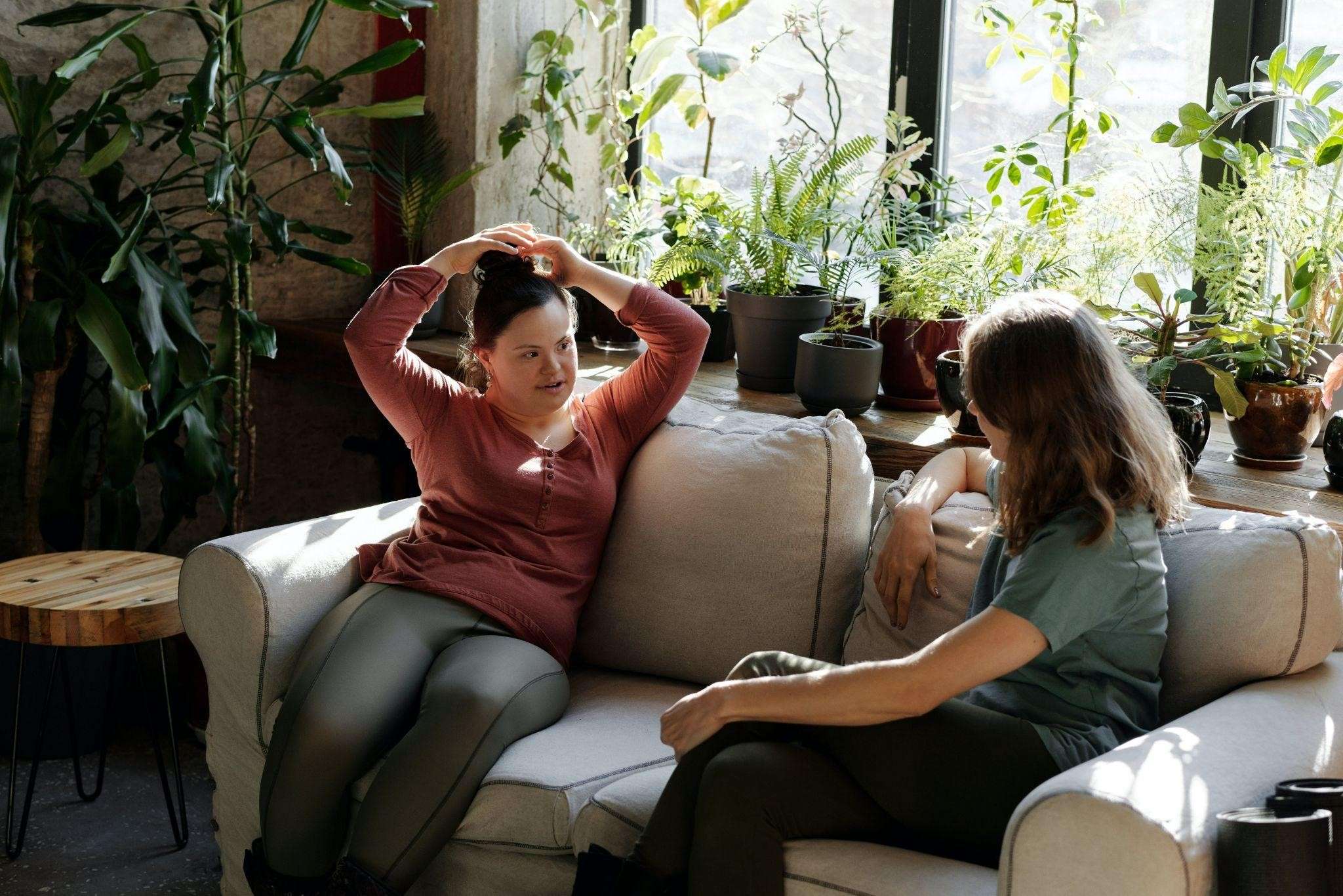Two women have a conversation on a couch surrounded by plants.